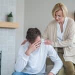 mother supporting her son who is sitting with his hand on his head, illustrating how to help a loved one with depression