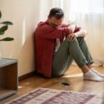 Depressed man sits on floor looking at mobile phone waiting for call from treatment center.