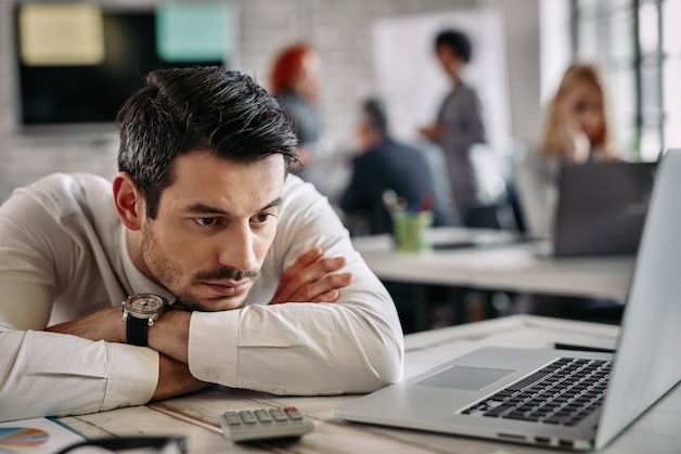 Young businessman thinking of something while leaning on his desk and using computer at work. There are people in the background.