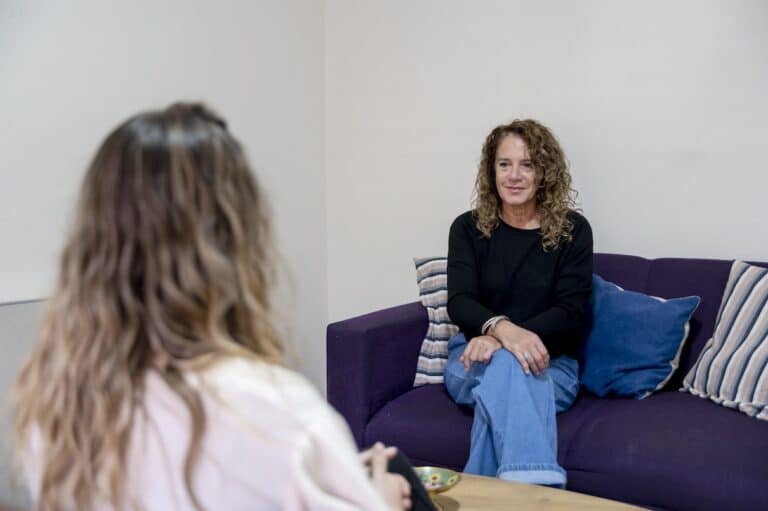 Woman sitting on purple sofa with legs crossed looking across the room at her therapist during outpatient mental health treatment