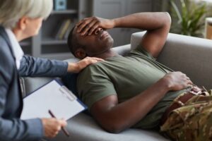 Man in military green tee with camo pants lying on a couch with a therapist next to him holding his shoulder