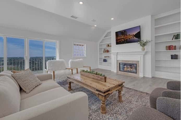 Bright modern A Mission for Michael living room with white built-in shelving, fireplace, rustic coffee table, and large windows.