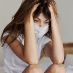 woman with hands on head and facemask on, wondering, "Is anxiety contagious?"
