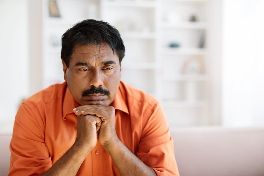 man in orange shirt, sitting pensively with hands under chin, experiencing existential anxiety