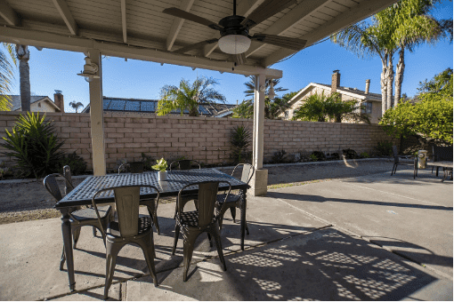 One of AMFM's covered outdoor patio facilities with a black metal dining table, chairs, ceiling fan, and view of a brick wall with palm trees beyond