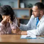 Crying woman sitting at wooden table with male doctor who is consoling her