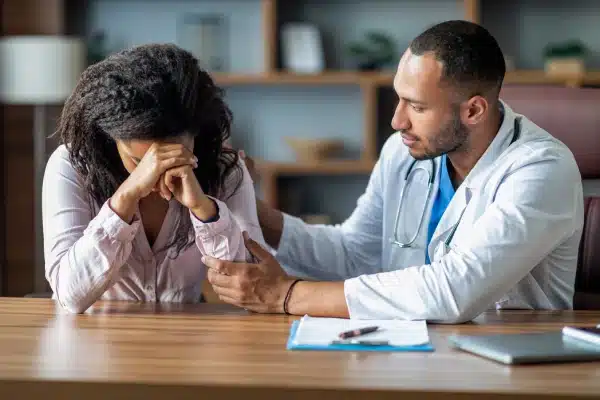 Crying woman sitting at wooden table with male doctor who is consoling her