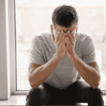 Man sitting on a window ledge with his head in his hands, asking is depression genetic?
