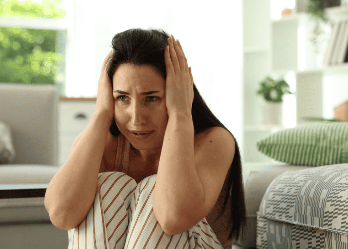 Woman sitting on the floor with her knees to her chest, her hands over her ears, with a panicked expression, in need of psychosis treatment