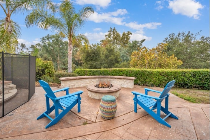 One of AMFM facilities with a backyard patio with a round stone fire pit, two bright blue Adirondack chairs, and a colorful woven stool.