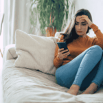 Woman sitting on the couch looking at her phone, struggling with social media and depression in adults