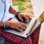 A young Black entrepreneur sits on a bench in a lively urban park, focused on his laptop while wearing headphones. The greenery and modern buildings provide a bright backdrop while he is searching bpd treatment cost