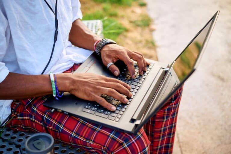 A young Black entrepreneur sits on a bench in a lively urban park, focused on his laptop while wearing headphones. The greenery and modern buildings provide a bright backdrop while he is searching bpd treatment cost