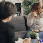 Woman crying and holding tissue to her nose as she sits on couch across from therapist