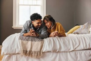 Couple laying on their stomachs leaning into eachother on a bed smiling