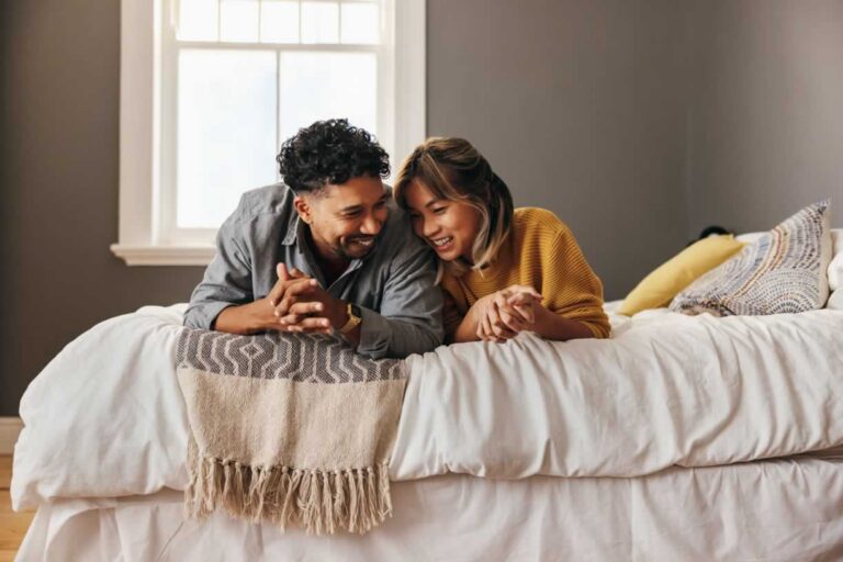 Couple laying on their stomachs leaning into eachother on a bed smiling