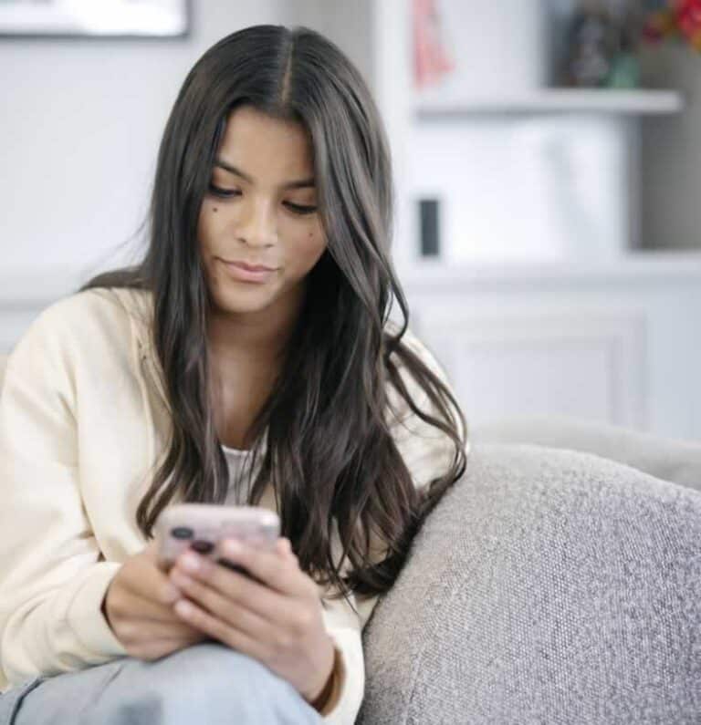 Young woman with long brown hair sitting on couch looking at her cell phone