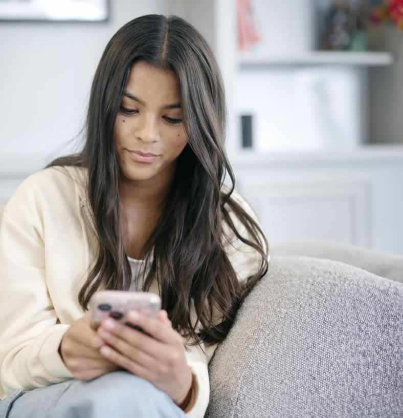Young woman with long brown hair sitting on couch looking at her cell phone