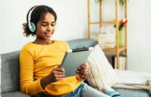 Young woman sitting on couch holding tablet and smiling as she looks at the screen and listens with headphones