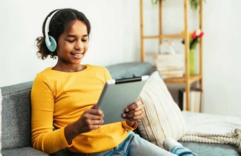 Young woman sitting on couch holding tablet and smiling as she looks at the screen and listens with headphones