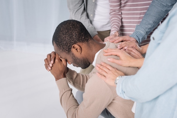 A small group of people standing with their hands on the back of one man who is hanging his head in his hands