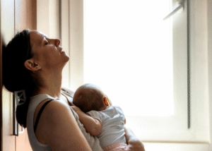 Woman sitting on the floor looking exhausted holding her baby, representing motherhood and mental illness