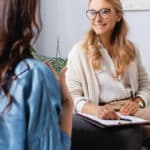 Blonde woman wearing glasses sitting on a chair across from another woman during therapy appointment