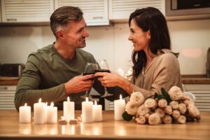 Man and woman looking at eachother as they cheers wine glasses among roses and multiple candles