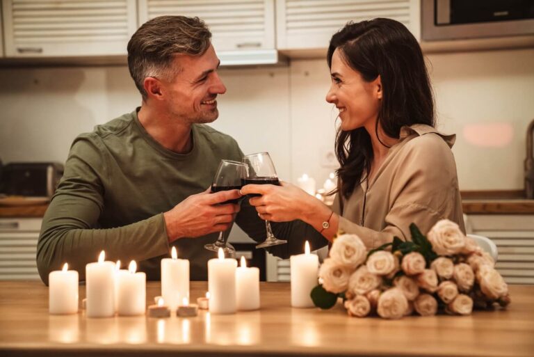 Man and woman looking at eachother as they cheers wine glasses among roses and multiple candles