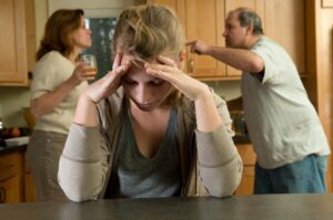 Teen girl holding her head in her hands on kitchen counter while her parents fight with each other in the background