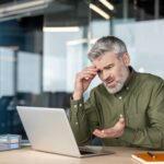 Man looking exasperated with hand on his head while sitting at a desk looking at his computer