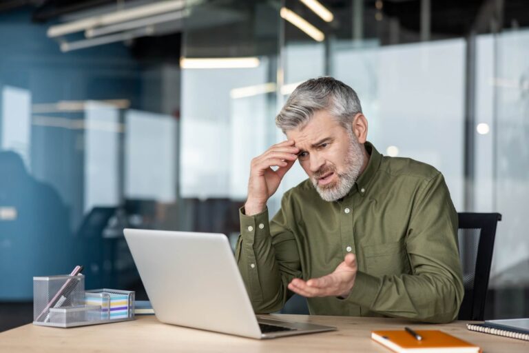 Man looking exasperated with hand on his head while sitting at a desk looking at his computer