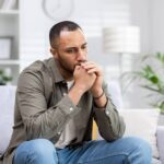 Man sitting on couch with a worried look on his face and hands held up covering his mouth as he thinks