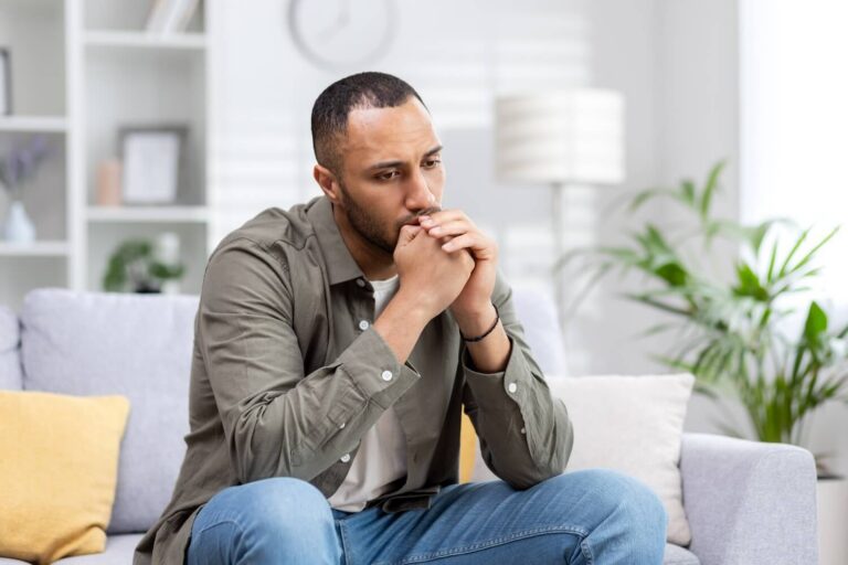 Man sitting on couch with a worried look on his face and hands held up covering his mouth as he thinks