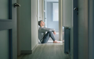 Man sitting on floor in hallway of home with head against the wall and eyes closed