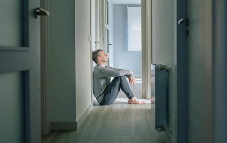 Man sitting on floor in hallway of home with head against the wall and eyes closed